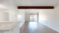 Located in Porter Ranch, Los Angeles, this 2-bedroom, 1-bathroom single-floor unit offers a practical and well-appointed layout. The open-plan living room features warm wood-look flooring, crown molding, an exposed wood ceiling beam, and a ceiling fan. A sliding glass door leads to a private outdoor balcony with open views of mature trees and the surrounding hillside landscape. The living area flows seamlessly into the kitchen, which is defined by rich cherry wood cabinetry, stainless steel appliances including a refrigerator and microwave, a white countertop peninsula, and a stainless steel sink — all finished with matching wood-look flooring. Both bedrooms are carpeted and bathed in natural light from large windows fitted with white horizontal blinds. The primary bedroom includes mirrored sliding closet doors that enhance the sense of space. The bathroom is clean and functional, featuring a full-sized soaking tub with an overhead shower, a window for natural light, a wood-toned vanity, and light stone-effect countertops. Clean neutral walls and white trim throughout the unit contribute to a bright, cohesive aesthetic.