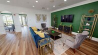 This single-floor space is thoughtfully designed with a cohesive contemporary aesthetic, uniting multiple functional areas through consistent design elements. Warm hardwood flooring flows throughout, complemented by crisp white walls, crown molding, and recessed lighting that collectively create a polished, inviting atmosphere.

The main living area serves as a focal point, anchored by a bold emerald green accent wall that houses a wall-mounted flat-screen television flanked by tall gold-framed shelving units and dark cabinet consoles. A plush navy blue sofa with yellow accent pillows faces the entertainment setup, centered around a dark wood coffee table and an ornate patterned area rug. Gold-accented glass side tables add a refined touch.

Adjacent to the living area, a sun-filled lounge nook features expansive arched windows on multiple walls, flooding the space with natural light. Blue upholstered accent chairs and an oval coffee table make this an ideal retreat for quiet conversation.

The dining area accommodates a large glass-top counter-height table surrounded by white upholstered chairs. A decorative starburst wall accent and colorful artwork add personality, while arched doorways connect the spaces seamlessly, reinforcing the open, flowing layout throughout.