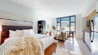 This single-floor studio is a well-appointed urban retreat that combines modern design with functional living. The open-plan layout seamlessly integrates the sleeping, living, and kitchen areas into one cohesive space, making excellent use of every square foot.

The main living area is anchored by large, floor-to-ceiling windows framed in sleek black metal, flooding the interior with abundant natural light and offering views of the surrounding cityscape. Light-toned hardwood flooring runs throughout, contributing to the bright and airy atmosphere. The sleeping area features a warm mid-century aesthetic, complemented by soft, neutral tones and stylish furnishings.

The kitchen is a standout feature, boasting warm wood upper cabinetry paired with clean white lower cabinets, a striking teal subway tile backsplash, light stone countertops, and a full suite of stainless steel appliances including a range, microwave, dishwasher, and refrigerator. A compact dining area sits adjacent, making the space ideal for everyday living and entertaining.

The bathroom is finished in a clean, minimalist style with a wide white vanity, undermount sink, and a glass-enclosed shower, all accented by crisp white surfaces and chrome hardware. Overall, this studio offers a modern, cohesive design with thoughtful details throughout.