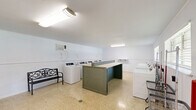 Located at 459 Apartment Court Dr in Baton Rouge, Louisiana, this shared laundry facility offers a well-organized, single-floor layout designed for functional, communal use. The room is finished with clean white walls accented by a horizontal rail detail and durable speckled terrazzo flooring throughout, providing a bright and easy-to-maintain environment.

The facility is equipped with a row of top-loading washing machines and matching dryers, arranged neatly along one wall for efficient use. A large central folding counter with a louvered base offers ample workspace for sorting and folding laundry, while wall-mounted shelving provides additional storage. Rolling wire laundry carts are conveniently available for transporting loads.

Natural light filters in through horizontal windows along one wall, complemented by overhead fluorescent lighting that keeps the space well-lit throughout the day. A wall-mounted air conditioning unit ensures comfort during Baton Rouge's warm seasons.

Additional amenities include a Coca-Cola vending machine and a snack vending machine, as well as a decorative bench seating area for waiting. A fire extinguisher is prominently mounted near the entry door, ensuring safety compliance. The space is accessible via a windowed exterior door, reinforcing its open and welcoming character. Overall, this laundry facility is a practical and well-equipped shared resource.