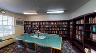The library at 2801 Montaluma Dr in Springfield, Illinois, is a single-floor space designed to serve as both a reading haven and a gathering area. The room is lined on multiple walls with tall, dark mahogany-finished bookshelves filled floor to ceiling with a diverse and extensive collection of books spanning a wide range of genres, offering ample reading options for visitors. At the center of the room sits a large rectangular table with a teal green surface, surrounded by cushioned chairs upholstered in a blue houndstooth pattern, making the space suitable for group reading sessions, meetings, or quiet study. The flooring features a patterned carpet with a rich floral motif in deep tones, adding warmth and texture to the room. Natural light filters in through multiple windows fitted with blinds, complemented by overhead fluorescent lighting that ensures the space remains well-lit throughout the day. Framed artwork adorns the walls, contributing a refined, welcoming atmosphere. A bulletin board near one window displays informational materials, adding a functional community element to the space. Additional freestanding bookshelves positioned near the entrance extend the collection into the adjoining corridor, seamlessly integrating the library with the broader facility.