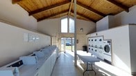 This single-story communal laundry facility is defined by its striking architectural design, most notably the soaring vaulted ceiling clad in warm natural wood planking with exposed beams that draw the eye upward and create an unexpectedly airy atmosphere. The peaked roofline is complemented by a large triangular window above the main entrance door, flooding the interior with generous natural light and offering views of the surrounding outdoor greenery.

The light-toned tile flooring runs throughout the space, enhancing the bright and clean aesthetic. Along the left wall, a continuous row of top-loading washing machines is neatly arranged, providing substantial laundry capacity. On the right side, stacked front-loading dryer units line the wall in organized columns, maximizing efficiency within the space.

A central structural support column anchors the room, while a long folding table positioned in the middle of the floor offers a practical surface for sorting and folding laundry. A wall-mounted air conditioning unit ensures year-round comfort. The overall ambiance is functional yet welcoming, with the warm wood ceiling providing a distinctive character not typically found in utility spaces of this kind.