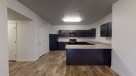 This single-floor unit offers a well-organized layout that balances open communal spaces with private retreats. The open-concept living and kitchen area forms the heart of the unit, where rich wood-look plank flooring flows throughout, grounding the space with a warm, cohesive aesthetic. The kitchen is outfitted with dark espresso cabinetry, granite-style countertops, and a full suite of black appliances including a refrigerator, range, over-the-range microwave, and dishwasher. A peninsula counter extends the workspace and provides additional seating or prep space, while upper and lower cabinets offer generous storage. The living area is open and airy, connected seamlessly to the kitchen and entry, creating an inviting, functional common space. Two bedrooms are finished with plush neutral carpeting and feature architectural shelf details near the ceiling, lending each room a distinctive character. Both bathrooms maintain a consistent design language, featuring dark vanity cabinetry with granite-style countertops, a full tub-shower combination, and the same wood-look flooring found throughout the rest of the unit. Clean white walls and neutral tones throughout create a bright, move-in ready environment that is both practical and visually cohesive.
