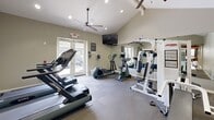 Located at 40800 Sunflower Rd in Murrieta, California, this fitness center offers a well-equipped exercise room designed to support a well-rounded workout routine. The space features vaulted ceilings that lend an open, airy feel, complemented by a ceiling fan and recessed lighting that ensure comfortable conditions during exercise. Natural light flows in through a set of glass double doors at the entrance, providing views of the surrounding exterior and keeping the space feeling bright and inviting.

The exercise room is outfitted with a selection of cardio equipment, including multiple treadmills and elliptical trainers, as well as a multi-station cable weight machine suitable for a variety of strength training exercises. Durable, interlocking tile flooring covers the workout area, offering both cushioning and easy maintenance. A wall-mounted flat-screen television is positioned for easy viewing during workouts. Mirrored wall panels enhance the sense of space while allowing users to monitor their form.

An adjoining restroom, accessible through a glass-paneled sliding door, adds convenience to the facility. Neutral-toned walls and functional furnishings throughout create a clean, focused environment. This fitness center provides residents with a practical and comfortable space for their daily exercise needs.