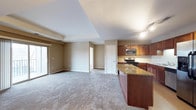 This single-floor residence offers a well-designed open-concept layout that seamlessly connects the kitchen and living areas. The kitchen is a standout feature, showcasing rich dark wood cabinetry, granite countertops with warm tonal variation, and stainless steel appliances including a range and built-in microwave. A peninsula-style countertop extends into the living space, providing a natural division while maintaining an open, airy feel. Track lighting overhead ensures the kitchen is well-lit for both cooking and entertaining. The adjoining living area is generously sized and finished with plush wall-to-wall carpeting in a neutral tone, creating a comfortable and inviting atmosphere. A tray ceiling detail adds architectural interest and a sense of height to the living space. A sliding glass door allows natural light to filter in and provides access to an outdoor area. The neutral color palette throughout — from the beige walls to the light carpet — offers a versatile backdrop that suits a variety of interior styles. Additional interior doors suggest further rooms beyond the main living space, completing this practical and cohesive single-level layout.