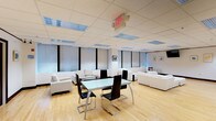 This single-floor 2-bedroom, 2-bathroom residence offers a well-organized layout designed for comfortable, modern living. The open-plan living and dining area serves as the heart of the home, showcasing light hardwood flooring that runs throughout and a dropped ceiling with recessed grid panels and integrated fluorescent lighting that ensures consistent, even illumination across the space. Large windows dressed with roller shades line the walls, allowing for ample natural light while maintaining privacy. The living area is furnished with multiple white upholstered sofas and seating arrangements, creating distinct lounging zones ideal for both relaxing and entertaining. A sleek glass-top dining table anchored by contemporary black cantilever chairs with chrome frames provides a stylish and functional dining space. The overall color palette is crisp and neutral — whites, blacks, and natural wood tones — lending the space a clean, minimalist character. Framed artwork on the walls adds subtle decorative accents without disrupting the understated elegance. The layout flows openly, giving the interior a spacious and airy feel. The two bedrooms and two bathrooms offer practical accommodation, completing this well-appointed and thoughtfully designed residence.