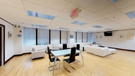 This single-floor 2-bedroom, 2-bathroom residence offers a well-organized layout designed for comfortable, modern living. The open-plan living and dining area serves as the heart of the home, showcasing light hardwood flooring that runs throughout and a dropped ceiling with recessed grid panels and integrated fluorescent lighting that ensures consistent, even illumination across the space. Large windows dressed with roller shades line the walls, allowing for ample natural light while maintaining privacy. The living area is furnished with multiple white upholstered sofas and seating arrangements, creating distinct lounging zones ideal for both relaxing and entertaining. A sleek glass-top dining table anchored by contemporary black cantilever chairs with chrome frames provides a stylish and functional dining space. The overall color palette is crisp and neutral — whites, blacks, and natural wood tones — lending the space a clean, minimalist character. Framed artwork on the walls adds subtle decorative accents without disrupting the understated elegance. The layout flows openly, giving the interior a spacious and airy feel. The two bedrooms and two bathrooms offer practical accommodation, completing this well-appointed and thoughtfully designed residence.