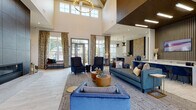 Located at 40000 Argento Way in Kannapolis, North Carolina, this single-floor clubhouse facility showcases a sophisticated and contemporary design that immediately impresses upon entry. The welcoming foyer greets visitors with tall ceilings, large windows dressed with white horizontal blinds, light-toned tile flooring, and carefully curated furnishings that set the tone for the entire space.

The expansive main lounge area is the architectural centerpiece of the building. Soaring vaulted ceilings — accentuated by a stunning wood-slat ceiling panel detail — rise dramatically overhead, while an oversized circular chandelier and modern linear pendant lighting illuminate the room. Floor-to-ceiling windows and multiple sets of glass-paned doors flood the space with natural light and offer views to the outdoors. Plush navy blue sofas and wingback chairs, warm golden ottomans, and layered area rugs create an inviting, upscale atmosphere.

A dramatic floor-to-ceiling dark tile fireplace wall serves as a bold focal point, anchoring one end of the lounge with a sleek linear fireplace and a raised concrete hearth. Adjacent to this is a resident lounge accessible through glass-paned barn-style doors. A staffed service counter and additional amenity areas are visible in the background, completing this well-appointed community gathering space.