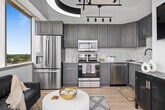 This single-story, one-bedroom, one-bathroom unit offers a thoughtfully designed layout that balances style and functionality. The open-concept kitchen is a standout feature, showcasing grey-toned cabinetry, stainless steel appliances including a range, microwave, and dishwasher, complemented by a light tile backsplash and white countertops. A dramatic black coffered ceiling with industrial-style track lighting adds a bold architectural contrast to the otherwise bright, neutral-toned space. Light wood-look flooring runs throughout the main living areas, enhancing the sense of openness and warmth.

The bedroom is serene and well-proportioned, featuring crisp white walls, the same light wood flooring, and ample space for a full furniture arrangement. Clean lines and neutral tones create a calm, restful atmosphere.

The bathroom features large-format hexagonal grey tile flooring, white walls, and a tub-shower combination with a curtain enclosure. A grey vanity with storage ties cohesively to the overall design palette.

The unit's layout, as seen in the 3D model, follows an angular configuration that maximizes the use of available square footage, providing a comfortable and functional living environment with a modern, cohesive aesthetic throughout.