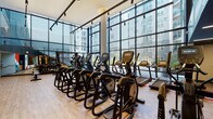 This expansive fitness center is thoughtfully designed to accommodate a wide range of workout styles across several well-defined zones. The cardio area is anchored by a striking wall of floor-to-ceiling glass windows that floods the space with natural light and offers outdoor views, creating an energizing atmosphere. Rows of treadmills, ellipticals, and stationary bikes from Matrix and other commercial-grade brands are arranged along the light-filled perimeter, all set on warm hardwood-style flooring accented by bold red stripe detailing.

The strength training zone features Matrix power racks, cable machines, adjustable benches, and a comprehensive free weight selection, complemented by large mirrors and vibrant, pop-art-style athletic murals that add a dynamic visual energy to the space. A dedicated functional training area is equipped with a heavy tire, plyo boxes, battle ropes, medicine balls, kettlebells, and a punching bag, illuminated by distinctive oversized pendant light fixtures that give this zone a boutique-gym aesthetic.

Linear LED lighting runs throughout the darker-ceilinged sections of the facility, providing consistent illumination across all areas. Glass partitions connect the various zones, maintaining an open, cohesive feel while defining distinct workout environments. Overall, the facility balances a modern industrial design with practical, professional-grade fitness amenities.