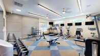Located at 400 Katrina Ct in Midlothian, VA, this fitness center offers a comprehensive workout environment within a single, open floor plan. The space is designed to accommodate a wide range of exercise needs, making it a functional and inviting facility for residents.

The room features a distinctive blue and tan checkered carpet tile flooring that adds a modern visual appeal underfoot. Overhead, ceiling fans with elegant brass fixtures provide air circulation, complemented by long linear LED light fixtures and recessed lighting that ensure the space is well-lit throughout.

Natural light filters in through multiple windows positioned along the walls, offering views of the surrounding outdoor environment. A row of flat-screen TVs is mounted along one wall, allowing users to stay entertained during cardio sessions.

The equipment layout is thoughtfully organized, featuring treadmills, elliptical trainers, upright and recumbent stationary bikes, a cable weight machine, adjustable weight benches, and a dumbbell rack. A large wall mirror enhances the sense of space and allows users to monitor their form during workouts.

Neutral-toned walls and a clean, uncluttered design contribute to a focused and motivating atmosphere. The fitness center is accessible via a keyed entry door and includes safety signage and a fire extinguisher, reflecting attention to safety and practicality.