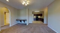 This single-floor space showcases an open-concept design that seamlessly connects the living area to the kitchen. The living room is finished with wall-to-wall carpeting in a neutral tone, creating a warm and comfortable atmosphere. A ceiling fan with a light fixture illuminates the space, while the textured ceiling adds subtle visual interest throughout.

A standout built-in feature is a wall-mounted desk or shelf nook with bracket supports, positioned along one wall of the living area. This practical element offers dedicated workspace or display space without occupying additional floor area.

The kitchen is accessible directly from the living space and is finished with rich, dark wood cabinetry that provides ample storage. Black appliances, including a range, microwave, and dishwasher, complement the cabinetry. A countertop with a sink lines the perimeter, and under-cabinet lighting enhances visibility for meal preparation. The kitchen flooring transitions from the carpeted living area to a smooth, hard surface.

The neutral beige walls throughout create a cohesive and adaptable backdrop, lending the space a clean, move-in-ready appearance well-suited for a variety of furnishing styles.