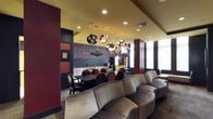 This single-floor space makes a striking first impression with its bold, eclectic interior design. The layout seamlessly blends a relaxed lounge area with a dedicated dining space, creating a versatile environment suited for both casual gatherings and more structured social events.

The lounge area is anchored by a generously sized curved sectional sofa upholstered in soft, neutral fabric, offering ample seating and an inviting atmosphere. Rich burgundy and dark wood accents — including statement columns and wall paneling — add warmth and visual depth throughout the space.

The dining area features a large dark wood table surrounded by upholstered chairs, set against a dramatic dark accent wall adorned with decorative script-style artwork in deep red tones. White cabinetry and light countertops along the back wall provide a bright contrast and suggest convenient service or kitchenette functionality.

One of the most captivating design elements is the collection of large, globe-shaped pendant lights suspended from the yellow-toned ceiling. These colorful, artistic fixtures cascade above the transition between the lounge and dining zones, serving as both functional lighting and sculptural focal points. Recessed lighting further enhances the overall illumination, while natural light filters in through windows along the perimeter, completing this richly layered and dynamic interior.