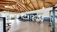 This fitness center offers a striking architectural backdrop for an effective workout experience. The defining feature of the space is its dramatic vaulted ceiling, showcasing exposed natural wood timber trusses and tongue-and-groove wood planking that rise to a generous peak, creating an open and airy atmosphere. Multiple ceiling fans are suspended from the trusses, ensuring comfortable air circulation throughout. Light-toned wood-style flooring runs across the entire floor, contributing to the bright and clean aesthetic. Large windows along the perimeter walls allow ample natural light to fill the room, while a glass door provides direct outdoor access and additional views to the exterior. A full-length mirror along one wall visually expands the space and supports strength training exercises. The fitness center is comprehensively equipped with a variety of cardio machines, including treadmills, stationary bikes, stair climbers, and ellipticals, as well as a range of strength and resistance machines, a cable multi-gym, and a freestanding pull-up and dip station. A dedicated dumbbell rack provides free weight options. Wall-mounted sconces add warm accent lighting, and a large flat-screen television is mounted on the wall for entertainment during workouts. The overall ambiance is bright, welcoming, and purpose-built for fitness.