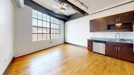 This sixth-floor loft unit in downtown Winston-Salem showcases an industrial-chic aesthetic with exposed ductwork, concrete columns, and track lighting on high ceilings. The open-concept main living area features warm hardwood floors and large factory-style windows that flood the space with natural light and offer urban views. The kitchen is equipped with stainless steel appliances, including a refrigerator, range, and dishwasher, complemented by dark wood cabinetry and granite countertops. The bathroom features large-format tile flooring, a granite-topped vanity with dark wood cabinetry, and a full mirror. A walk-in closet with wire shelving provides practical storage.