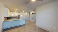 This single-floor residence showcases a contemporary open-concept design with clean lines and modern finishes throughout. The kitchen serves as a focal point, featuring crisp white cabinetry extending to the ceiling, a bold hexagonal tile backsplash, and a full suite of stainless steel appliances including a refrigerator, range, microwave, and dishwasher. A peninsula-style island with a light countertop and undermount sink provides additional workspace and casual seating, accented by stylish pendant lighting overhead. The kitchen flows seamlessly into the living area, where wood-look luxury vinyl flooring continues underfoot and a ceiling fan provides comfort and circulation. A distinctive sliding barn door adds architectural character, offering a stylish division between spaces. The bedroom is finished with plush carpeting, a ceiling fan, and a generously sized window with blinds that brings in natural light and outdoor views. The bathroom features a dual-sink vanity with white cabinetry, light countertops, wide mirrors, and wood-look flooring, complemented by a tiled shower surround. Recessed lighting throughout the unit ensures consistent, well-distributed illumination, while the neutral color palette creates a cohesive and inviting atmosphere across all spaces.