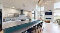 Located at 5532 38th St S in Fargo, ND, this thoughtfully designed community amenity space seamlessly combines a full kitchen, lounge, and game room into one expansive, single-floor layout. The architecture is defined by dramatic vaulted ceilings with exposed wood beam trusses, flooding the space with a sense of grandeur and openness. Herringbone-pattern hardwood flooring flows through the kitchen and lounge areas, adding warmth and visual interest throughout. The fully equipped kitchen features a bold teal island with dark stone countertops and bar seating, white shaker cabinetry, a patterned tile backsplash, a stainless range hood, and black appliances, all framed by an industrial-style steel and glass partition wall. The lounge area is anchored by a floor-to-ceiling stacked stone fireplace topped with a large-screen TV, flanked by oversized windows that draw in abundant natural light and offer views of the surrounding landscape. The game room and lounge area feature carpeted flooring, a full-size pool table, large communal dining tables, high-top seating, and a bank of windows that brighten the space throughout the day. Together, these zones create a versatile, welcoming environment ideal for gathering, relaxing, and recreation.