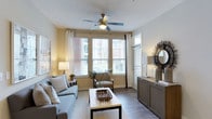 This single-floor residence offers a welcoming and well-appointed living area designed with both comfort and functionality in mind. The living room serves as the focal point of the space, bathed in abundant natural light streaming through multiple large windows dressed with white curtains and adjustable blinds. These windows offer views of the surrounding exterior, enhancing the sense of connection to the outdoors.

The room features warm, hardwood-style flooring that runs throughout, providing a clean and cohesive aesthetic. A ceiling fan with integrated lighting is centrally mounted, ensuring air circulation and supplemental illumination. Soft yellow-toned walls contribute to the inviting atmosphere, complementing the neutral furnishings that include a gray sofa, a wooden coffee table, and a coordinating accent chair.

A dark wood storage credenza is positioned along one wall, styled with decorative accessories including a sunburst mirror, adding visual interest and practical storage. The overall ambiance is calm and contemporary, with a balanced mix of natural textures and modern finishes that create a comfortable and livable environment.