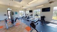 Located in Mansfield, TX, this single-story fitness center offers a comprehensive workout facility designed to support a wide range of exercise routines. The space is organized into distinct workout zones, each purpose-built for specific training needs. The cardio area is outfitted with multiple treadmills, elliptical machines, and rowing machines, all positioned along large windows that frame views of the surrounding green landscape, allowing natural light to fill the room. Ceiling fans and overhead lighting maintain a comfortable and well-lit environment throughout.

A dedicated strength training section features cable machines and selectorized equipment, complemented by open floor space accented with orange accent tiles on a durable rubberized floor. Wall-mounted flat-screen televisions are positioned throughout the facility for entertainment during workouts. A separate spin studio houses a row of stationary cycling bikes alongside stability balls and other functional fitness accessories stored on a dedicated rack. A water fountain is conveniently accessible within the space. The facility's white walls, elevated ceilings, and multi-pane windows create a bright, airy atmosphere, while direct access to the outdoors adds convenience and connectivity to the exterior surroundings.