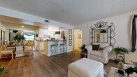 This single-floor residence offers a thoughtfully designed open-concept layout that seamlessly connects the living, dining, and kitchen areas. The living room features warm hardwood-style flooring, a textured ceiling, and large windows dressed with elegant draping, allowing generous natural light to filter in. A sliding glass door opens to a private patio, further enhancing the sense of openness. An ornate iron mirror and framed wall art add character to the space.

The kitchen is equipped with white cabinetry, white appliances including a range and refrigerator, and granite-style countertops. A peninsula with bar seating provides a casual dining option, while a separate dining area accommodates formal meals. The kitchen opens directly to the living and dining spaces, promoting a connected and social atmosphere.

The primary bedroom features plush carpeting, a ceiling fan, natural light from a window with blinds, and ample space for a full furniture suite. It connects to a generous walk-in closet with wire shelving. A second bedroom also offers carpeted flooring and a sliding closet. The bathroom includes a vanity with stone-style countertops, a tub-shower combination, and hardwood-style flooring, completing this comfortable and functional residence.