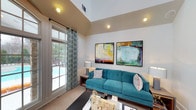 This well-appointed single-floor space makes a strong first impression with its dramatic vaulted ceilings that add an expansive, airy quality to the interior. The living area is the centerpiece of the space, designed with a contemporary aesthetic that balances comfort and style. A striking teal tufted sofa anchors the room, complemented by a neutral area rug and a sleek coffee table that together create an inviting gathering area. Two large, vibrant abstract paintings adorn the walls, adding bold splashes of color that enliven the otherwise understated palette of warm whites and soft neutrals. Recessed lighting throughout the ceiling ensures the space is well-lit, while a large window fitted with elegant patterned drapes floods the room with abundant natural light. The window also frames a view of an outdoor pool area, bringing a resort-like ambiance to the interior. The vaulted ceiling design, with its elevated upper section, further enhances the sense of openness and grandeur. Overall, the space offers a thoughtfully curated environment that feels both sophisticated and welcoming, making it well-suited for relaxed everyday living or casual entertaining.
