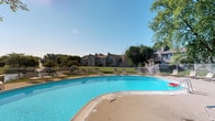 Crooked Creek offers a standout outdoor amenity area centered around a large, freeform swimming pool with clear blue water. The pool deck is finished in a neutral concrete surround and is equipped with multiple lounge chairs, providing ample space for relaxation under open skies. A white metal security fence encloses the pool area, ensuring both safety and a clean, defined perimeter. Beyond the fence, a broad stretch of well-kept green lawn extends toward the residential buildings, which feature traditional architectural elements including pitched rooflines, chimneys, and balconies. Mature trees frame the edges of the property, offering natural shade and a sense of established landscaping. The overall setting is bright and open, benefiting from abundant natural light and clear sightlines across the grounds. The combination of the expansive pool, manicured lawn, and classic building architecture creates a cohesive and inviting outdoor environment well-suited for warm-weather enjoyment. The single-level layout of the community contributes to an accessible and approachable atmosphere throughout the property.
