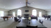 This single-story space showcases an open-concept layout beneath striking vaulted ceilings that lend a sense of grandeur and airiness to the interior. The architecture is highlighted by tall, arched windows that flood the room with natural light, framing the neutral gray walls and creating a bright, welcoming atmosphere. Ceiling fans with recessed lighting are positioned along the ridge of the vaulted ceiling, ensuring both comfort and even illumination throughout.

A central fireplace with a white mantel serves as the focal point of the main seating area, flanked by a wall-mounted flat-screen television above it. Dark leather upholstered sofas and accent chairs are arranged around the fireplace, creating a comfortable lounge area. A large dark-stained dining table with matching chairs occupies the foreground, offering a dedicated space for dining or group activities.

To one side, a billiard table is visible, adding a recreational element to the space. The flooring transitions between hard surface and carpet, defining different functional zones. Overall, the space presents a versatile, social environment suited for gatherings, relaxation, and entertainment.