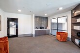 Located at 13200 133rd Ct in Overland Park, Kansas, this contemporary single-floor office space is thoughtfully designed to balance productivity and comfort. The layout seamlessly integrates multiple functional zones, each finished with modern materials and purposeful design elements throughout.

The primary workroom features a long, dark-toned conference-style desk lined with multiple computer workstations, accompanied by sleek ergonomic task chairs. A sculptural pendant light fixture anchors the space from above, while multiple windows frame views of the surrounding neighborhood, flooding the room with natural light and creating an open, airy atmosphere.

A welcoming lounge area provides a relaxed counterpart to the workstations, highlighted by a bold tufted orange velvet accent chair positioned beside a floor-to-ceiling built-in dark wood bookcase with integrated accent lighting. This area also includes a semi-private pod seating unit, ideal for focused work or informal meetings.

A dedicated service counter features a clean white quartz surface set against a striking geometric 3D tile accent wall, paired with warm wood cabinetry below — offering a refined, functional workspace for administrative tasks.

Throughout, the space is unified by patterned carpet flooring, neutral white walls, and recessed lighting, contributing to a professional yet inviting environment well-suited for a leasing office or business service center.
