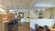 Located at 4130 Times Square Blvd in Dublin, Ohio, this two-floor commercial space combines classic design sensibilities with a warm, welcoming atmosphere throughout. The main level features rich hardwood flooring that runs seamlessly across an open layout, anchored by a white beadboard reception or service counter with a dark countertop. Pendant lighting hangs from the ceiling, complementing the crown molding and neutral, textured wall treatments that lend the space a refined character. A large ornate mirror in a decorative wood frame adds depth and elegance to the main area, while bar-height seating along a wooden counter provides a functional yet stylish touch. Mirrored wall panels near the service area further enhance the sense of openness and light. The lounge area, likely situated on a lower level, offers a cozy and intimate setting with plush leather club chairs, a round wooden coffee table, and soft table lamp lighting. Wall-mounted televisions are present in multiple areas, catering to guest comfort and engagement. Wainscoting details and consistent neutral tones tie the two levels together, creating a cohesive aesthetic. The overall ambiance is polished and comfortable, well-suited for a professional service environment focused on hospitality and client experience.
