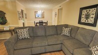 This single-floor space showcases an open-concept layout that seamlessly connects the living room and dining area, creating a cohesive and functional flow throughout. The living room is anchored by a large, plush gray sectional sofa with a chaise, offering ample seating and a comfortable focal point for the space. Decorative patterned throw pillows add a subtle contrast to the neutral upholstery. Warm beige walls paired with light hardwood flooring contribute to an inviting atmosphere throughout.

Crown molding runs along the upper perimeter of the walls, adding a touch of classic architectural character. A textured ceiling with visible HVAC vents indicates practical climate control infrastructure. Wall art featuring ornate ironwork-style pieces adds decorative depth to the living area.

The adjacent dining area is defined by a dark wood table and matching chairs, centered beneath an elegant crystal chandelier that serves as a statement lighting fixture. A framed mirror on the dining room wall reflects light and enhances the sense of openness. A doorway visible from this vantage point suggests additional rooms beyond, contributing to the overall functional layout of the space.