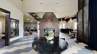 Located at 10800 McCombs St in El Paso, Texas, this single-floor community clubhouse offers a generously sized, open living space designed with a modern yet inviting aesthetic. The interior is immediately distinguished by its bold geometric hexagonal tile flooring in a gray and cream chevron pattern, which flows seamlessly throughout the entire space.

A dramatic double-sided mosaic tile fireplace serves as the architectural centerpiece, clad in rich brown and copper mosaic tiles that rise toward the soaring ceiling. Exposed dark wood ceiling beams add warmth and contrast against the neutral toned walls, while multiple pendant lights and recessed fixtures provide layered illumination throughout.

The living area is thoughtfully divided into distinct zones, including a lounge seating area with plush sofas and armchairs, a bar counter with high stools, and a computer workstation area. Large sliding glass doors and oversized windows invite abundant natural light and offer views of the surrounding exterior. Dramatic floor-to-ceiling drapes in deep navy add a refined touch to select corners of the space.

A welcoming entry corridor features the same signature tile flooring and leads visitors into the main gathering area. Interior windows with dark trim frames connect adjacent spaces, maintaining visual openness while defining functional zones. Overall, the space exudes a sophisticated, contemporary clubhouse ambiance well-suited for community gatherings and social interaction.