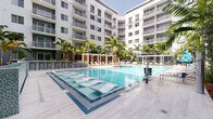 This amenity space is defined by its resort-inspired design, centered around a generously sized swimming pool with crystal-clear blue water and integrated in-pool lounge chairs that allow residents to relax partially submerged. The pool deck is finished in light grey porcelain tile, creating a clean, contemporary aesthetic that extends throughout the courtyard. Tropical palms and richly planted garden beds line the perimeter, adding vibrant greenery and a lush, warm-climate ambiance to the space.

A poolside bar with counter seating provides a social gathering point, while shaded pergola cabanas feature horizontal wood slat screens and cushioned chaise lounges, offering a comfortable retreat from the sun. The surrounding multi-story building wraps around the courtyard, with large glass sliding doors connecting interior amenity spaces directly to the pool deck, enhancing indoor-outdoor flow.

Ground-level walkways bordered by tropical plantings, including palms, bromeliads, and ornamental grasses, provide pleasant circulation throughout the space. Glass-panel railings maintain open sightlines while delineating different zones. Overall, the space delivers a polished, modern tropical atmosphere well-suited for leisure and relaxation.
