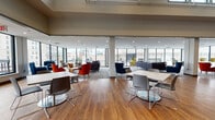 Located at 10 Chestnut St in downtown Springfield, Massachusetts, this single-floor space is designed to support a dynamic, collaborative environment. The open-plan layout is defined by warm hardwood flooring, recessed lighting, and expansive floor-to-ceiling windows that flood the interior with natural light while offering unobstructed views of the surrounding urban cityscape.

The space is thoughtfully divided into distinct functional zones. A lounge area features an eclectic mix of seating — including bold red and navy accent chairs, deep sectional sofas, and a distinctive olive wingback chair — arranged around a central coffee table atop patterned carpet tiles, creating an inviting, comfortable atmosphere. Nearby, multiple white-topped tables with contemporary chrome-legged chairs provide flexible workstation and dining arrangements suited for individual use or group collaboration.

A separate, more enclosed area offers additional white tables and grey chairs, accented by colorful artwork on the walls, and benefits from elevated ceiling height for a spacious feel. Adjacent to this, a counter area with warm wood cabinetry and a row of orange bar stools presents a casual dining or service station setup. Throughout the space, glazed partition walls and glass entry doors maintain visual connectivity while defining separate zones, reinforcing the space's open and transparent architectural character.