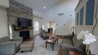 This single-floor space is defined by its dramatic vaulted ceilings in the main living area, which lend an open, airy quality to the room. The focal point of the living space is a floor-to-ceiling stacked stone fireplace, paired with a mounted flat-screen television above a wooden mantle, creating a warm and stylish centerpiece. Hardwood flooring runs throughout, providing a cohesive and elegant foundation. The living area is comfortably furnished with a sectional sofa, patterned accent chairs, an ottoman, and a central coffee table, all arranged on a large area rug. Natural light filters in through multiple windows fitted with blinds. Recessed lighting in the ceiling supplements the ambient light effectively. An open doorway connects the living area to an adjoining room, visible in the background, which houses a grand piano, suggesting a dedicated music or entertainment space. The overall ambiance is warm and welcoming, combining classic architectural details like the stone fireplace with a neutral, contemporary color palette that makes the space feel both functional and refined.