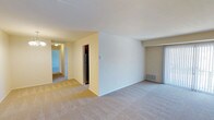 This single-floor residential unit offers a clean and functional layout designed for comfortable everyday living. The main living area is spacious and features wall-to-wall carpeting in a neutral beige tone, creating a warm and inviting atmosphere throughout. The open floor plan allows for a natural flow between the living space and the adjacent kitchen area, which is partially visible and includes a stainless steel refrigerator. A central dividing wall helps define the distinct zones within the unit while maintaining an open feel. To one side, a sliding glass door provides access to the outdoors and allows natural light to filter into the living area. A hallway leads from the main living space to additional rooms beyond, offering a sense of depth and privacy within the unit. Walls are painted in soft, neutral tones that complement the light carpeting throughout. Practical features include visible floor vents for climate control and a thermostat on the central wall. The overall ambiance is clean, bright, and move-in ready, with a straightforward layout that provides both functionality and versatility for a variety of living arrangements.