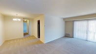 This single-floor residential unit offers a clean and functional layout designed for comfortable everyday living. The main living area is spacious and features wall-to-wall carpeting in a neutral beige tone, creating a warm and inviting atmosphere throughout. The open floor plan allows for a natural flow between the living space and the adjacent kitchen area, which is partially visible and includes a stainless steel refrigerator. A central dividing wall helps define the distinct zones within the unit while maintaining an open feel. To one side, a sliding glass door provides access to the outdoors and allows natural light to filter into the living area. A hallway leads from the main living space to additional rooms beyond, offering a sense of depth and privacy within the unit. Walls are painted in soft, neutral tones that complement the light carpeting throughout. Practical features include visible floor vents for climate control and a thermostat on the central wall. The overall ambiance is clean, bright, and move-in ready, with a straightforward layout that provides both functionality and versatility for a variety of living arrangements.