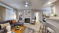 This single-floor residence offers a well-organized layout across two bedrooms, one bathroom, a living room, a combined kitchen and dining area, a dedicated office, and a connecting hallway. The living room serves as the heart of the home, featuring a tiled fireplace with brass-accented doors, crown molding, carpet flooring, and a ceiling fan with light fixture. Natural light enters through a large window dressed with blinds and drape panels. The combined kitchen and dining area includes warm wood cabinetry, tile flooring, and white appliances, opening toward the dining space, which features a mirrored wall accent that visually expands the room. The primary bedroom includes a closet and a window providing natural light. The bathroom offers tile flooring, a vanity with white cabinetry, a large mirror, recessed lighting, and a tub-shower combination. The dedicated office provides a quiet, functional workspace with crown molding and carpet flooring. Throughout the residence, a consistent warm gray color palette, crown molding, and carpet in main living areas create a cohesive and polished ambiance.