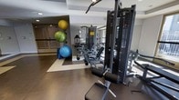 This fitness center is designed to support a comprehensive workout experience within a single-level layout. The space features a thoughtfully organized floor plan with distinct zones for different types of training. A multi-station cable and weight machine anchors the strength training area, accompanied by a variety of resistance equipment and adjustable benches. The flexibility and functional training zone is stocked with stability balls in multiple sizes, yoga mats, and other accessories neatly arranged along the wall. Large mirrors line the walls, enhancing the sense of space and allowing users to monitor their form during exercise. The flooring transitions between durable dark rubber and lighter-toned sections, clearly delineating workout zones while providing cushioning and traction. Natural light filters in through windows, contributing to a bright and energizing atmosphere. The overall aesthetic is modern and clean, with a neutral color palette of grey and white that keeps the environment focused and professional. The space is well-maintained and efficiently arranged to maximize usability, making it suitable for a full range of fitness routines from strength training and stretching to functional movement work.