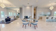 Located at 14770 SW 264th St in Homestead, FL, this single-floor space offers a thoughtfully designed open-plan layout that seamlessly connects its key functional areas. Throughout, large-format polished porcelain tile flooring reflects natural light, contributing to a bright and airy atmosphere.

The kitchen is a standout feature, showcasing high-gloss cabinetry in a soft gray tone, a sleek white countertop island with seating for multiple guests, and a linear glass tile backsplash. A dramatic tiered crystal chandelier serves as a striking focal point overhead, while a window provides natural light and outdoor views.

The living area exudes sophistication with soaring ceilings, full-length wall mirrors that amplify the sense of space, and a generous arrangement of seating in neutral and navy tones. Multiple windows frame lush outdoor greenery, flooding the room with natural light. A second crystal chandelier with tray ceiling detailing further elevates the refined aesthetic.

A dedicated workstation area features built-in desk surfaces with multiple computer stations, tall windows overlooking outdoor landscaping, and ample room for focused productivity. A utility room provides additional built-in cabinetry with a tile backsplash, offering organized storage and workspace support.

Overall, the space presents a clean, contemporary aesthetic with luxurious finishing touches throughout.