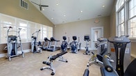 Located at 193 Tenby Chase Drive in Delran, New Jersey, this single-floor fitness center offers a well-equipped workout facility designed to support a comprehensive exercise routine. The space features soaring vaulted ceilings accented by a ceiling fan, creating an open and airy atmosphere that enhances comfort during workouts. Recessed lighting throughout ensures the room is consistently well-lit, supplemented by natural light streaming in through multiple windows along one wall. The fitness center is outfitted with a diverse array of Cybex cardio machines, including treadmills, elliptical trainers, and an upright bike, catering to a variety of cardiovascular training preferences. Strength training is supported by a multi-station cable machine, a free-weight bench, and additional resistance equipment. The neutral-toned walls and light-colored flooring contribute to a clean, modern aesthetic that keeps the focus on functionality. A dedicated doorway leads to an adjacent room, offering additional utility to the space. The overall layout is thoughtfully organized to allow for comfortable movement between equipment, making this fitness center a practical and inviting destination for daily exercise.