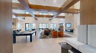 This generously sized single-floor community space is designed with a contemporary aesthetic that balances sophistication and comfort. The most striking architectural feature is the dramatically high ceiling articulated by a bold grid of exposed wooden beams, which adds warmth and structural character to the otherwise crisp white interior. Large floor-to-ceiling black-framed windows and glass doors line the perimeter, flooding the space with natural light and offering clear views of a lush outdoor patio and pool area. Light-toned porcelain tile flooring runs throughout, unifying the various functional zones seamlessly.

The open-plan living area serves as the social centerpiece, furnished with plush sectional sofas, curved burnt-orange accent chairs, and a round wooden coffee table arranged around a sophisticated area rug. A dramatic floor-to-ceiling stone and wood fireplace surround anchors one wall, topped by a wall-mounted television. Adjacent to this lounge area, a pool table provides recreational entertainment.

The kitchen area features dark cabinetry with white quartz countertops, a decorative light-blue tile backsplash, built-in appliances, and a long bar-height counter with seating. Pendant and chandelier lighting throughout enhance the polished, modern ambiance of the entire space.