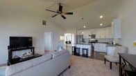 This single-floor residence is defined by its open-concept layout and striking vaulted ceilings that lend an expansive, airy quality to the main living area. A ceiling fan is centrally positioned overhead, enhancing both airflow and visual appeal throughout the space.

The living room is anchored by a generously sized light-gray sofa and complemented by soft, neutral-toned carpeting that adds warmth underfoot. Recessed lighting and a floor lamp provide layered illumination, while a wall-mounted television rounds out the comfortable, functional setup.

Flowing seamlessly from the living area is the kitchen, which features crisp white cabinetry, stainless steel appliances including a range and over-the-range microwave, and a kitchen island with bar-height seating. The island serves as both a prep space and a casual dining area, with dark bar stools adding a stylish contrast to the light cabinetry. The kitchen floor transitions to hardwood-style flooring, clearly delineating it from the carpeted living zone.

Adjacent to the kitchen, a defined dining area is visible, offering a dedicated space for more formal meals. The open sightlines throughout the main floor create a cohesive, connected atmosphere ideal for both everyday living and entertaining. The neutral color palette and clean architectural lines give the entire space a modern yet inviting character.