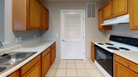 This single-floor residence offers a practical and cohesive layout with a neutral color palette throughout. The kitchen is efficiently designed in a galley-style configuration, featuring warm honey-toned oak cabinetry, light countertops, tile flooring, and a full suite of appliances including an electric range and refrigerator. A louvered interior door adds a classic touch and leads to an adjacent utility area. The dining area connects openly to the kitchen and is anchored by a stylish tiered pendant light fixture, with vaulted ceilings adding a sense of airiness to the space. Tile flooring continues seamlessly through this area before transitioning to plush wall-to-wall carpeting in the living and bedroom areas. The living room is generously proportioned, featuring soaring vaulted ceilings, a sliding glass door that invites natural light, and an open feel enhanced by neutral beige tones. Multiple bedrooms offer comfortable, carpeted spaces with large windows dressed in blinds that allow for ample natural light. Two full bathrooms are finished with tile flooring, white tub-shower combinations, and warm wood vanity cabinetry, providing clean and functional spaces throughout the home.