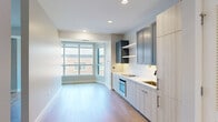 Located at 1201 North Clark Street in Chicago's vibrant Gold Coast neighborhood, this one-bedroom, one-bathroom unit offers a well-designed layout across a single floor. Light hardwood flooring runs throughout, complementing the neutral color palette and recessed lighting that keep the space feeling bright and airy.

The open-concept kitchen and living area serves as the heart of the unit. The kitchen features two-tone cabinetry — pairing light wood lower cabinets with slate-blue upper cabinets and open shelving — alongside a white quartz countertop, a patterned tile backsplash, a built-in oven, cooktop, and a deep sink. Large windows at the far end of the living area flood the space with natural light and provide views of the surrounding cityscape.

The bedroom is generously sized, with its own large corner window that continues the theme of natural light. The unit also includes two closets for ample storage and a dedicated laundry area. A hallway connects the main living spaces to the private areas of the unit.

The bathroom is finished with clean, modern details, including large-format white tile flooring, a wood-toned vanity with a white countertop and brushed nickel hardware, a full soaking tub with a tiled surround featuring an accent stripe, and a well-lit vanity mirror.