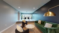 This well-appointed single-floor amenity space combines functionality and modern design throughout its thoughtfully arranged areas. The entry lobby greets visitors with light wood-look flooring, crisp white trim, elegant pendant chandeliers, and a built-in mailbox bank — setting a clean, contemporary tone from the moment of arrival. Large glass doors at the entrance allow natural light to pour in, creating an airy and open feel.

Adjacent to the lobby, the club room serves as the social heart of the space. A lounge area anchors one side, featuring a tufted green velvet sofa set against a dramatic dark accent wall, complemented by a natural fiber rug and a round marble-top coffee table. The room transitions via a raised platform step into a dedicated dining area, where a long communal table with sleek black chairs is positioned directly in front of expansive floor-to-ceiling sliding glass doors. These doors offer unobstructed views of the greenery outside, flooding the area with natural light.

A separate, quieter co-working room provides a focused environment for productivity, outfitted with paired white desks, warm wood accent chairs, light gray walls, and tasteful botanical artwork. Light wood flooring flows consistently throughout the entire space, unifying each area into a cohesive and polished whole.