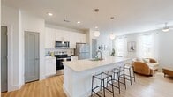 This thoughtfully designed one-bedroom, one-bathroom residence offers a bright and modern living experience across a single floor. The open-concept layout seamlessly connects the kitchen and living room, creating an inviting and functional space. The kitchen is a standout feature, showcasing white shaker cabinetry, light quartz countertops, a tiled backsplash, and a full suite of stainless steel appliances including a range, over-the-range microwave, and refrigerator. A large center island with bar seating and globe pendant lighting serves as both a prep space and casual dining area. The living room benefits from abundant natural light streaming through multiple windows dressed with sheer white curtains, complemented by light hardwood-style flooring that runs throughout the unit. The bedroom is a comfortable retreat, featuring a ceiling fan, dual windows with outdoor views, and a generous closet with double doors. The bathroom is finished in crisp white tones with a white vanity, quartz countertop, large mirror with globe sconce lighting, a tub-shower combination with tile surround, and convenient access to a walk-in closet with built-in shelving. Clean lines, neutral tones, and consistent modern finishes give this residence a cohesive and polished aesthetic throughout.