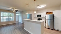 This single-floor residence offers a thoughtfully designed open-concept layout that seamlessly connects the kitchen, dining, and living areas. The kitchen is a standout feature, outfitted with crisp white shaker-style cabinetry, a classic white subway tile backsplash, light quartz countertops, and a full suite of stainless steel appliances including a refrigerator, electric range, and built-in microwave. A charming built-in wine rack adds a distinctive decorative touch. A central kitchen island with pendant lighting serves as an elegant focal point, providing additional prep space and casual seating opportunities. The open living area is finished with durable hardwood-style flooring and features a ceiling fan with light kit, creating a bright and airy atmosphere. The bedroom is generously proportioned, featuring soft carpeting underfoot, dual windows that invite ample natural light, and a ceiling fan for year-round comfort. The bathrooms are cleanly finished with white vanity cabinetry, quartz countertops, and light wood-look flooring. One bathroom includes a tiled tub-shower combination, while another features a compact vanity with a window for natural light. Neutral tones and consistent finishes throughout lend the space a cohesive, modern feel.