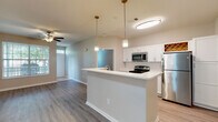 This single-floor residence offers a thoughtfully designed open-concept layout that seamlessly connects the kitchen, dining, and living areas. The kitchen is a standout feature, outfitted with crisp white shaker-style cabinetry, a classic white subway tile backsplash, light quartz countertops, and a full suite of stainless steel appliances including a refrigerator, electric range, and built-in microwave. A charming built-in wine rack adds a distinctive decorative touch. A central kitchen island with pendant lighting serves as an elegant focal point, providing additional prep space and casual seating opportunities. The open living area is finished with durable hardwood-style flooring and features a ceiling fan with light kit, creating a bright and airy atmosphere. The bedroom is generously proportioned, featuring soft carpeting underfoot, dual windows that invite ample natural light, and a ceiling fan for year-round comfort. The bathrooms are cleanly finished with white vanity cabinetry, quartz countertops, and light wood-look flooring. One bathroom includes a tiled tub-shower combination, while another features a compact vanity with a window for natural light. Neutral tones and consistent finishes throughout lend the space a cohesive, modern feel.