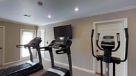 This single-floor fitness space is designed to provide a functional and focused workout environment. The room features neutral-toned walls with crisp white crown molding and baseboards, contributing to a clean and orderly atmosphere. Recessed ceiling lighting ensures the space is well-lit throughout, creating a bright and energizing setting for exercise.

The room is equipped with multiple treadmills and an elliptical trainer, offering a variety of cardio workout options. A wall-mounted flat-screen television is positioned at eye level, allowing users to stay entertained or catch up on news during their workouts. The carpeted flooring provides a degree of comfort underfoot, while the overall layout allows for easy movement between the machines.

A standard doorway visible along the wall suggests connectivity to an adjacent room or corridor, adding convenience for users moving through the facility. A window near the entrance allows for a modest amount of natural light to filter into the space, enhancing the overall ambiance. The room's straightforward, no-frills design keeps the focus on fitness, making it a practical and accessible facility for everyday use.