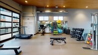 Located at 1513 W Meeker St in Kent, Washington, this fitness center is designed to support a comprehensive workout experience within a single, open-plan space. The room is distinguished by its warm wood-planked ceiling, recessed lighting, and expansive floor-to-ceiling windows that flood the interior with natural light and offer views of the surrounding neighborhood. The durable rubberized flooring provides a practical, cushioned surface throughout.

Cardio equipment, including multiple treadmills, an elliptical, and a rowing machine, is positioned to take full advantage of the panoramic windows. Full-length mirrors line one wall, visually expanding the space and allowing users to monitor their form. A dedicated strength training area includes a multi-function cable/smith machine, a dumbbell rack with a full range of weights, and an adjustable workout bench. A storage rack holds stability balls, medicine balls, and resistance accessories for functional and group training.

Clerestory windows provide additional light along the upper walls, while athletic silhouette murals add a motivating, energetic character to the space. The overall ambiance is modern, bright, and well-organized, creating an inviting environment suited for a wide range of fitness activities.